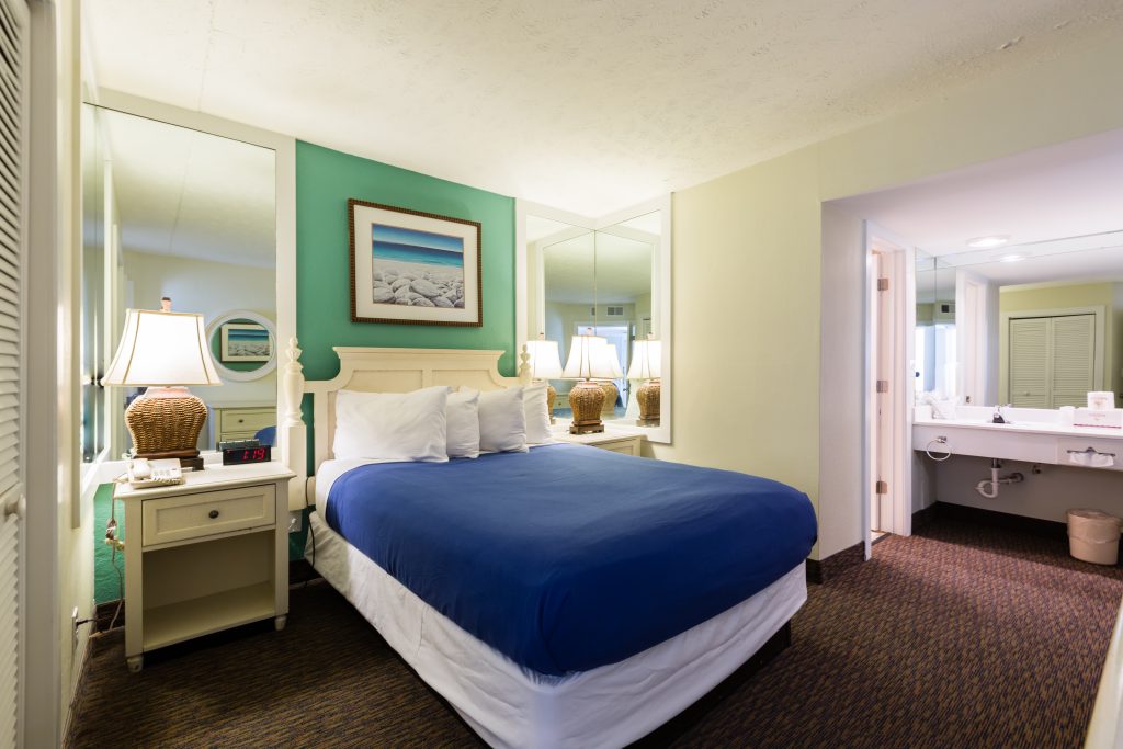 A hotel room with a king-size bed, nightstand, lamp, mirror, painting, and bathroom with sink.