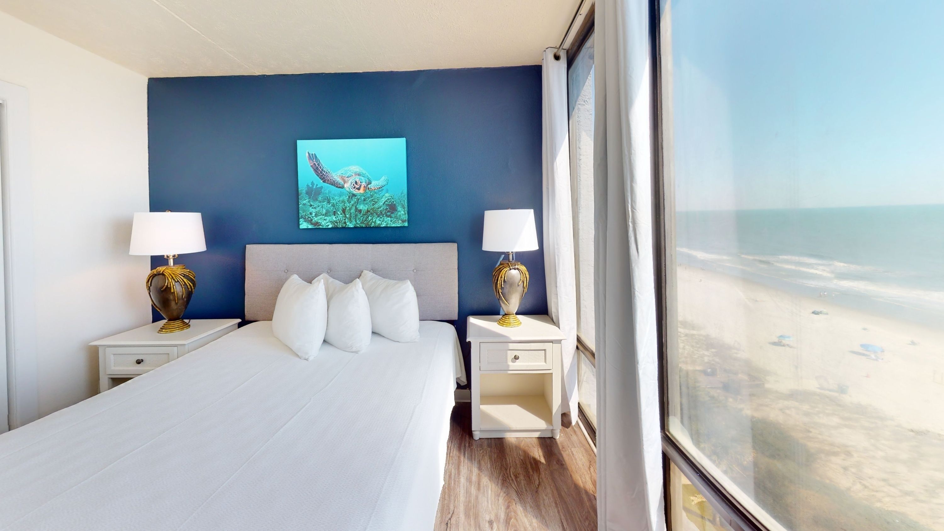 A wide view of a hotel room with a bed, two nightstands, and a large window showing a beach and ocean view.
