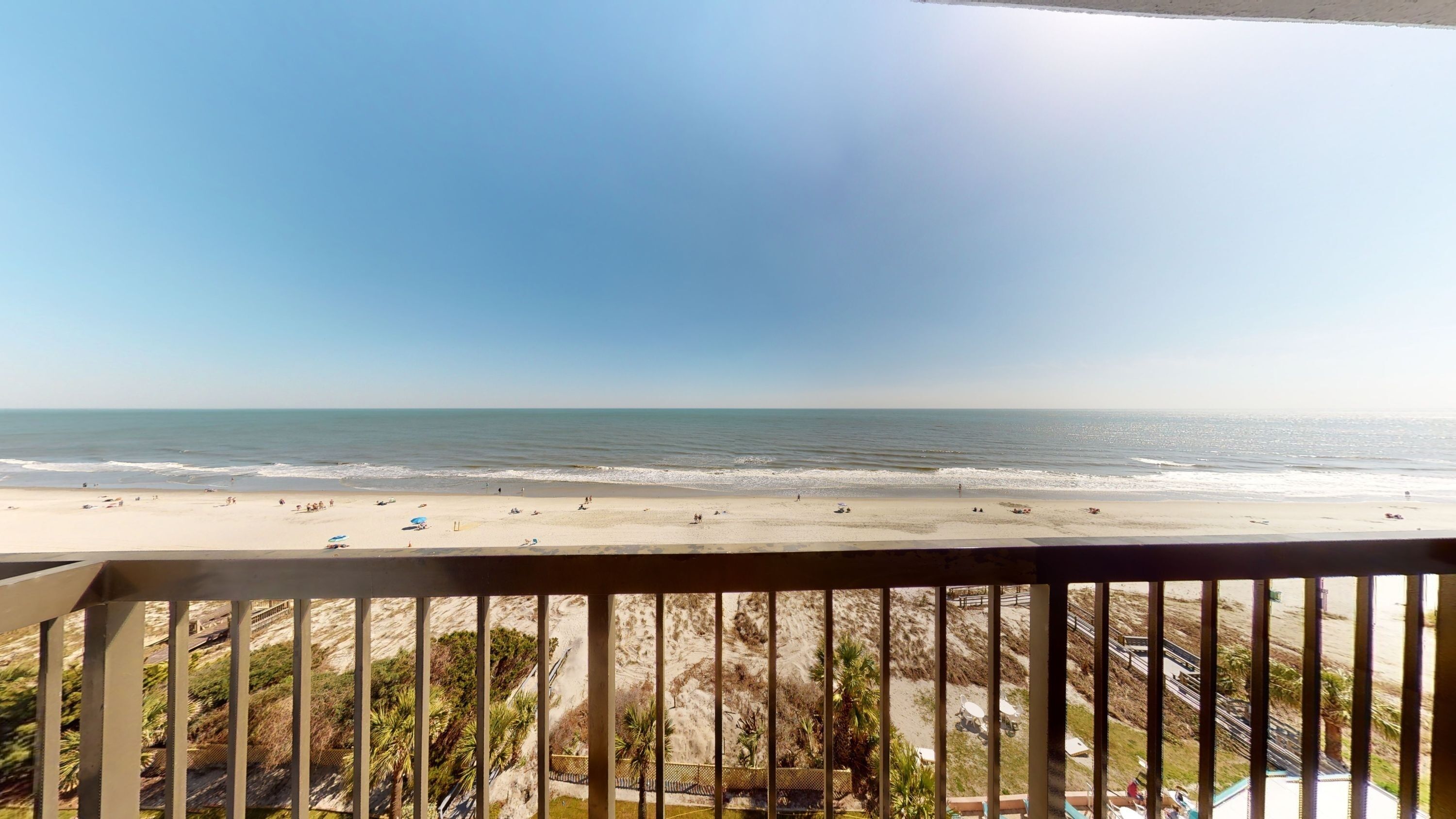 A beach view from a balcony with a railing, overlooking the ocean and sandy beach.
